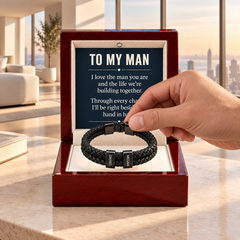 Hand lifting a black double-strand braided personalized bracelet with DANIEL and MEGAN name beads from a red luxury gift box with To My Man card